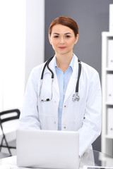 Happy doctor woman at work. Portrait of female physician using laptop computer while standing near reception desk at clinic or emergency hospital. Medicine concept