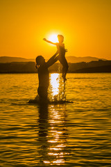 Silhouettes of father throwing his child in water on sunset. Family playing in the sea. Sky is yellow, reflects in water. Mountain ridge in background. Copy space, design, background, meme, wallpaper.