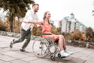 Good speed. Young man having fun and smiling while being in the park with disabled girlfriend and pushing her wheelchair