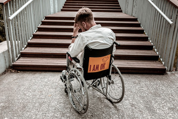 Big barrier. Sad disabled young man closing his eyes while sitting in front of the stairs without...