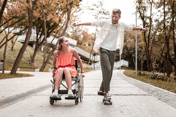 Riding skateboard. Cheerful emotional young man putting hands up and riding a skateboard while spending time with his cute disabled girlfriend