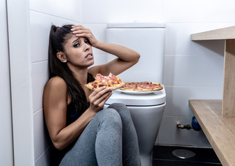 Young beautiful bulimic woman sitting on the bathroom floor eating pizza looking guilty