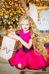Fototapeta na wymiar Cute little girl with long curly blond hair at home near a Christmas tree with gifts and garlands and a decorated fireplace 