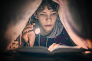 portrait of young teenager reading a book with a light lying under the blanket at night f