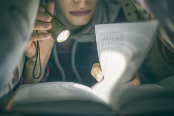 portrait of young teenager reading a book with a light lying under the blanket at night f