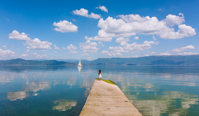 Little girl looking at a sailboat on the edge of a lake.  Standing on concrete bridge, back turned. Clouds and mountains reflecting in water. Blue. White clouds.  Landscape. Nature. Magical. Summer. - Powered by Adobe