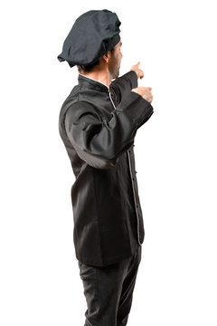 Chef Man In Black Uniform Pointing Back With The Index Finger Presenting A Product From Behind On Isolated White Background