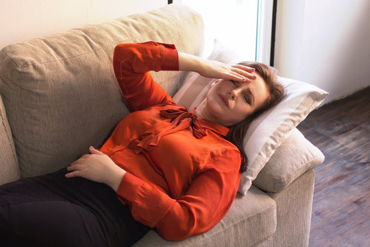 Sick Woman With Headache Laying On Couch. Stressed Sick Young Businesswoman Resting On Sofa With Her Hand At Her Head And Sad Emotions On Her Face.