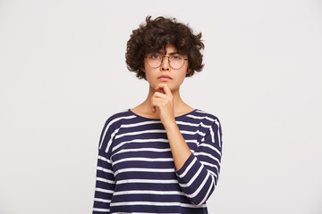 Fototapeta na wymiar Studio shot of thoughtful girl ponders on some idea, keeps one hand on chin wears white and blue stripped sweatshirt and transparent black-framed round glasses isolated over white background