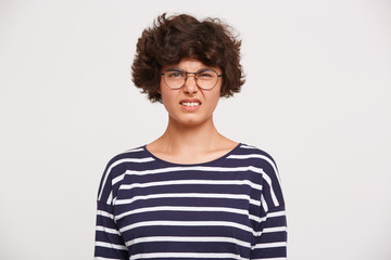 Portrait of young woman with curly brown hair, wears white and blue stripped sweatshirt and transparent black-framed round glasses feels disgust wrinkles nose isolated over white background
