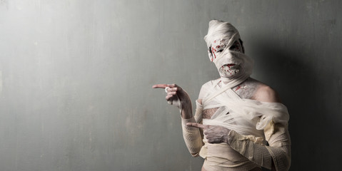 Terrorific mummy pointing to the lateral on textured wall background.  Halloween holidays