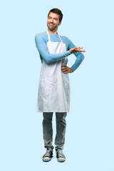 Full body of Man wearing an apron presenting a product or an idea while looking smiling towards on blue background