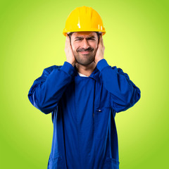 Young workman with helmet covering both ears with hands. Frustrated expression on green background