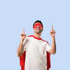 Fototapeta premium Superhero man with mask and red cape pointing with the index finger a great idea and looking up on isolated blue background