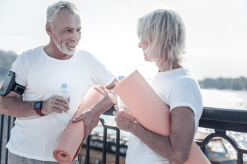 Fototapeta premium Healthy people. Pleased bearded male person holding bottle in right hand and having case for telephone on arm, looking at his wife