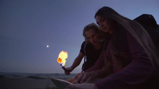 Young couple using a flaming torch to read a map at dusk in the wilderness during a hiking trip with backpacks.