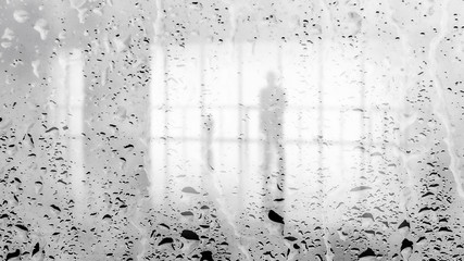 Blurred silhouette of man in the light hall through large wet glass wall. Black and white image