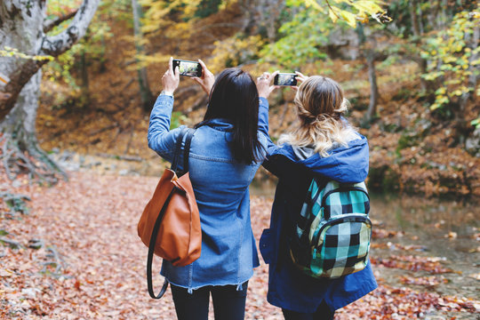 Two young girl friends walking in an autumn forest and taking pictures of the beautiful nature on the smartphone. Walk in the woods. Girlsfriends hiking at fall.