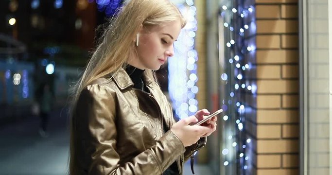 SLOW MOTION: Woman is window shopping at night. Young blonde girl is passing shop window display downtown, while typing on phone. writes a message about discounts. Portrait shot. wireless earphones