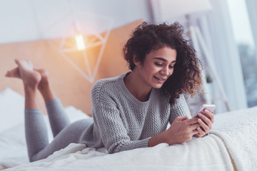 Relaxed woman. Relaxed young woman feeling relieved while lying in bed holding her phone in her...