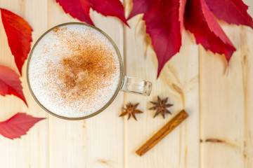 Obraz na płótnie Canvas Hot drink, coffee latte or cappucino with cinnamon on milk foam in a glass mug, spices and red autumn leaves on wooden background, cold season concept, top wiew