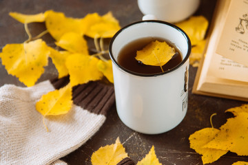 two white cups with tea in yellow leaves with a book and a knitted scarf, the concept of warmth and autumn sadness