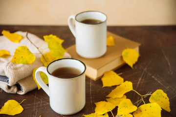 Naklejka na ściany i meble mugs surrounded by yellow foliage with a book and a knitted scarf, the concept of warmth and autumn sadness
