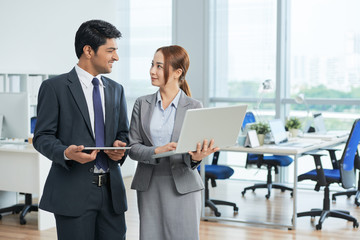 Young businesswoman using laptop standing with businessman using tablet pc, they talking at modern office