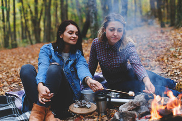 Fototapeta na wymiar Two young girls girlfriends roasting sweet marshmallow on a fire in the evening in the autumn forest.