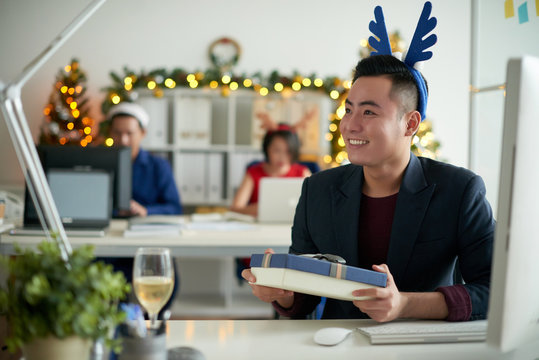 Happy Excited Young Asian Guy In Blue Antler Headband Sitting At Table With Computer And Receiving Christmas Gift From Colleague In Modern Office