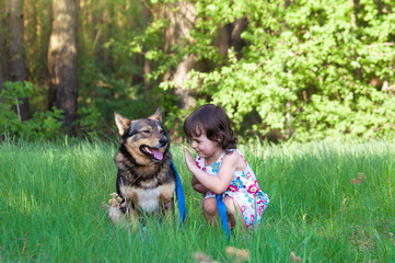 Naklejka na ściany i meble Happy little girl with the dog sits on the green lawn