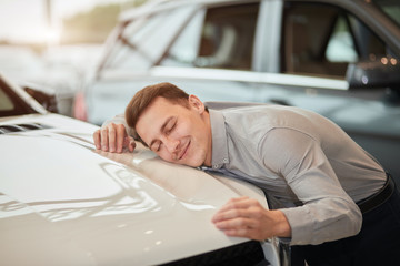 Close up of skillful male sales rep hugging a new car with closed eyes at dealership, displays all...