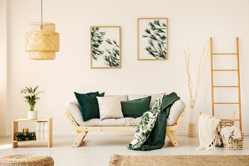 Bright living room interior with simple posters on wall, sofa with cushions and two blankets and wooden end table with books and fresh plant in real photo - obrazy, fototapety, plakaty