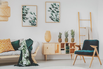 White living room interior with retro armchair, wooden cupboard with books, straw lamp and three pineapples, posters on wall and couch with blankets in the real photo
