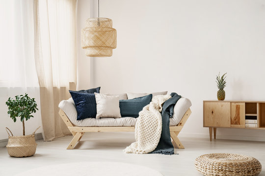 Light lounge with pillows and two blankets placed in white sitting room interior with window with curtains, straw lamp and fresh plant on floor in real photo