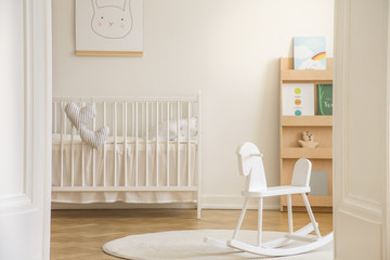 Rocking horse on rug in white kid's bedroom interior with rabbit poster above cradle. Real photo