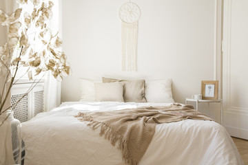 Blanket on white bed with cushions in minimal bedroom interior with plant and table. Real photo - obrazy, fototapety, plakaty