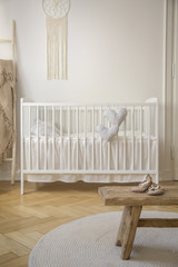 Wooden stool with shoes on round rug in bright baby's bedroom interior with white cradle. Real photo