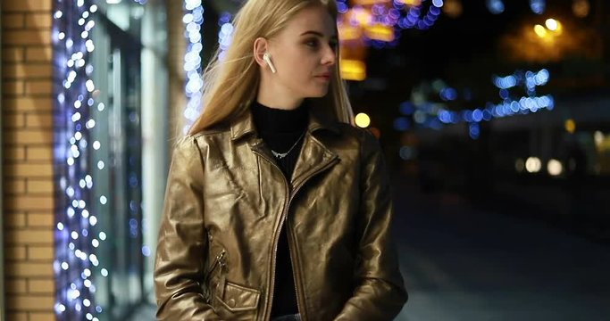 Attractive long-haired blonde young caucsian girl happily smiles straight to camera. City lights on the background. Positive mood, being happy, cheerfulness. Female portrait. wireless earphones