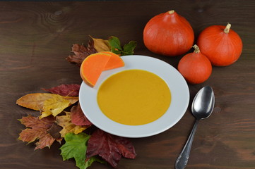 autumnal pumpkin soup in a bowl with hokkaido pumpkins, leaves and a spoon on a brown wooden table