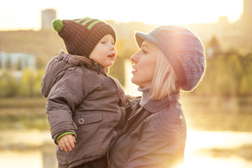 Mother in a warm jacket and knitted hat hugging her baby boy walking in the autumn park. stylish...