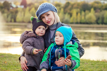 happy and beautiful blonde woman in hat with two baby son eating a chocolate bar in a warm jacket in the autumn park against the backdrop of the lake. stylish mother with child tasty snack picnic