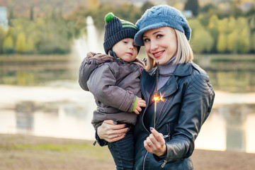 Fototapeta premium Mother in a warm jacket and knitted hat hugging her baby boy walking in the autumn park. stylish blondewoman with a child against the backdrop of the lake
