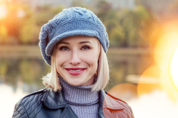 Young blonde happy smiling woman in beige coat and warm hat outdoor closeup portrait in the autumn park. Cold outside. Winter spring clothes