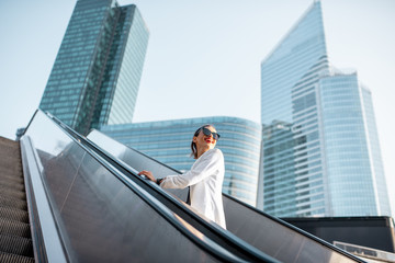 Stylish businesswoman in white suit going up on the escalator at the business centre outdoors with skyscrapers on the background in Paris