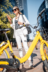 Fototapeta na wymiar Portrait of a stylish business woman in white suit standing with bicycle and phone at the financial district with modern buildings on the background