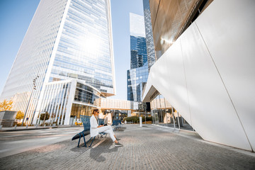 Young business woman in white suit sitting on the bench outdoors at the modern financial district. Wide angle view