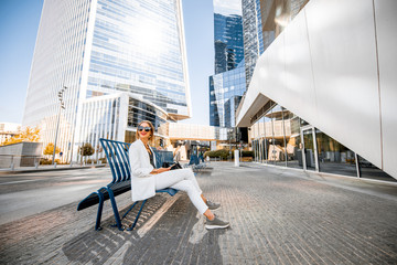 Young business woman in white suit sitting on the bench outdoors at the modern financial district