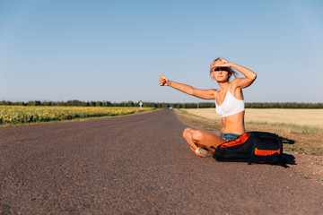 A girl with blond hair with a backpack sits on an asphalt road and catches a car