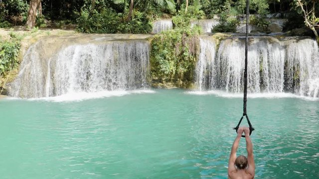 Young caucasian man with blond hair rope swinging into turquoise natural pool at Cambugahay Falls in Siquijor Island, the Philippines in ultra slow motion